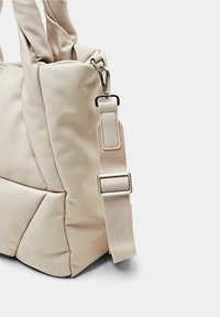 Beige mulepose med en blød, tekstureret yderside. Har tykke stofhåndtag og en aftagelig, justerbar rem med metalbeslag.