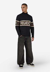 Marineblauer Rollkragenpullover mit cremefarbenem und braunem "PEGADO"-Band, gerippten Bündchen und Saum, kombiniert mit dunklen, weit geschnittenen Jeans und schwarzen Turnschuhen.