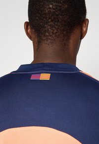 Achteraanzicht van een persoon in een marineblauwe en perzikkleurige sportshirt met een klein rechthoekig logo in rood, blauw en geel gestreept nabij de kraag.