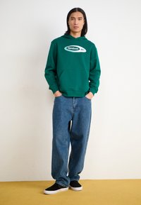 Hanorac verde cu glugă, având un mare logo alb Reebok. Asortat cu blugi albaștri, largi, și pantofi negri fără șireturi. Fundal simplu.