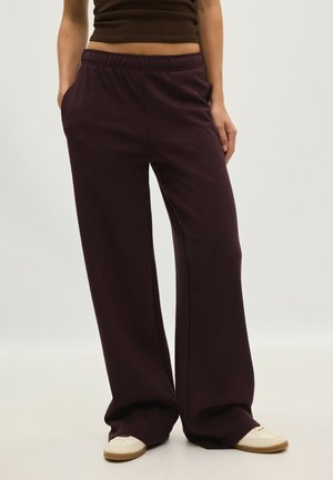 Personne portant un pantalon ample marron foncé avec une taille élastique et des baskets blanches, debout les mains dans les poches sur un fond neutre.