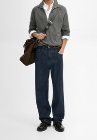 Grijze ritstrui, witte overhemd met kraag, donkerblauwe ruime jeans, bruine lederen riem, zwarte schoenen, bruine tas met schouderband.