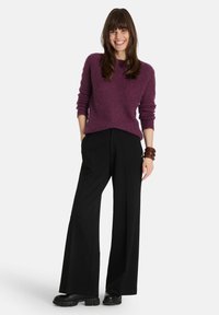 Lila Strickpullover mit rundem Ausschnitt, kombiniert mit hochtaillierten schwarzen weit geschnittenen Hosen. Das Model trägt klobige schwarze Schuhe und ein braunes Armband.