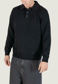 Maglione polo nero lavorato a maglia con colletto e polsini a costine, caratterizzato da un'apertura con tre bottoni e una vestibilità rilassata, abbinato a pantaloni in tessuto grigi.