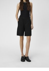 Haut noir sans manches à texture côtelée, associé à un short noir sur mesure avec poches latérales et un design lisse et structuré. Chaussures noires à enfiler.