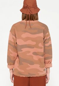 Felpa mimetica marrone con maniche arrotolate e orlo sfilacciato, abbinata a un cappello a secchiello marrone coordinato con un laccio sul retro.