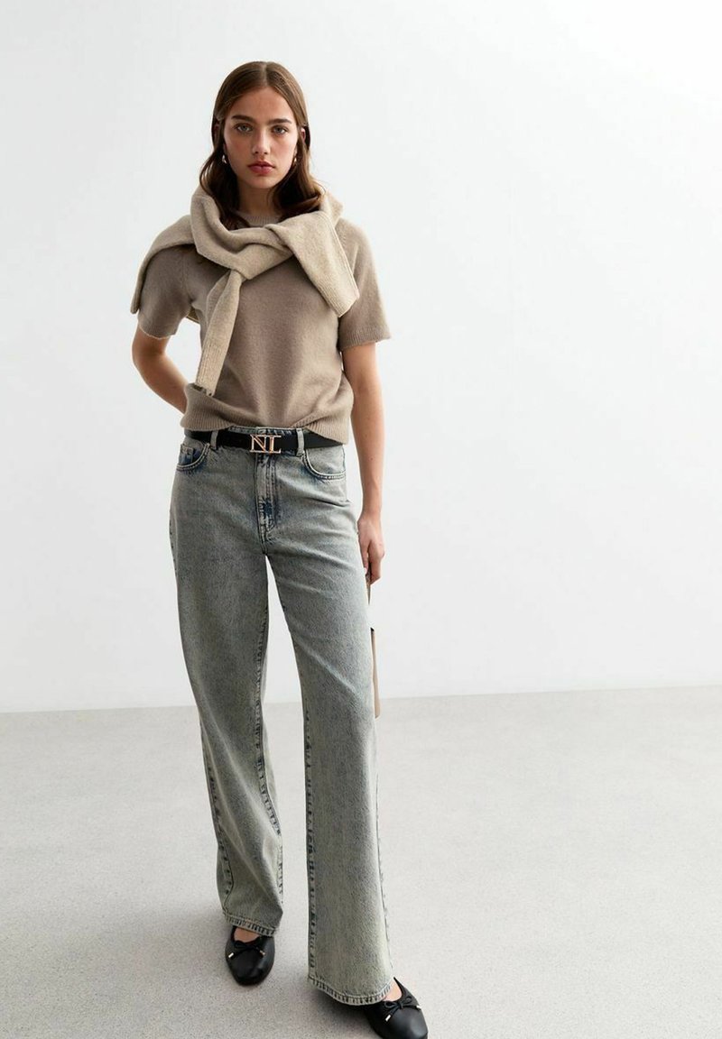 Beige gebreide trui over de schouders, gecombineerd met lichtgewicht jeans met wijd benen. Zwarte riem met gouden hardware. Zwarte platte schoenen. Eenvormige achtergrond.