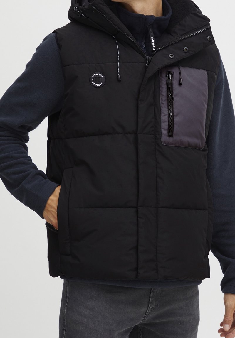 Homme portant un gilet sans manches noir matelassé avec une poche poitrine zippée et une capuche, par-dessus une chemise à manches longues sombre et un jean gris.