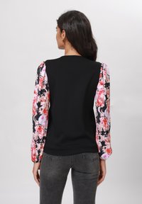 Svart topp med blomsterprintede ermer i rosa og svart, med puffede skuldre og en figursydd kropp, kombinert med grå jeans.