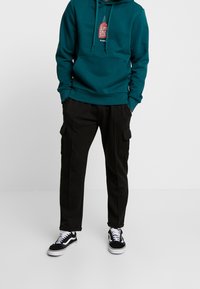 Felpa con cappuccio turchese con grafica frontale, abbinata a pantaloni cargo neri con tasche laterali e sneakers bianche con dettagli neri.