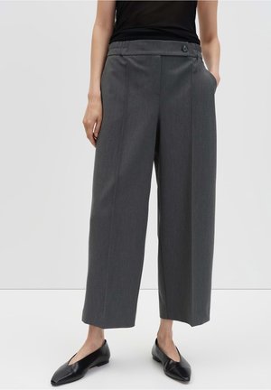 Kobieta w szarych, szerokich spodniach z wysokim stanem, z czarnym guzikiem i czarnych, spiczastych balerinach, stojąca na jednolitym tle.