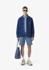 Niebieska dżinsowa kurtka z przednimi kieszeniami, jasnoniebieska koszulka polo, pasujące szorty, wzorzysta torba na ramię oraz beżowe sneakersy. Modelka w okularach przeciwsłonecznych.