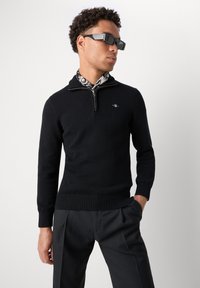 Hombre con suéter negro de media cremallera, bufanda estampada, pantalones negros y gafas de sol rectangulares oscuras, de pie sobre un fondo blanco liso.