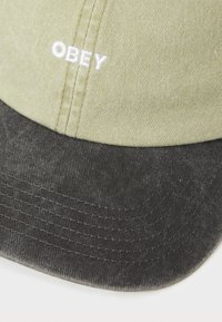 Katoenen pet met een lichtgroene bovenkant en een donkergrijze klep. Voorzien van witte borduursel "OBEY" aan de voorkant en een gestructureerde stof.