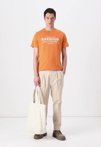 T-shirt de algodão laranja com texto branco, calças plissadas bege, botas castanhas e uma tote bag de lona branca. Conjunto simples e casual.