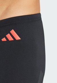 Pantalones cortos deportivos negros con una textura suave; cuenta con un logo rojo de adidas en el lateral y un pequeño acento reflectante en el borde inferior.
