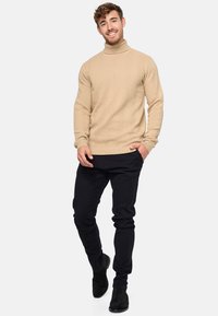 Pleteni pulover s visokim ovratnikom u bež boji s teksturiranim cik-cak uzorkom, u kombinaciji s crnim slim-fit hlačama i crnim čizmama do gležnja.