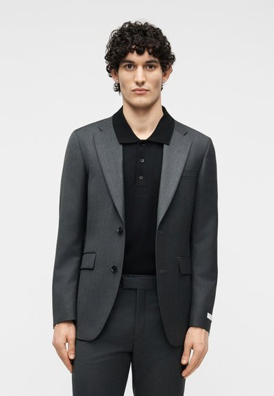 Νέος άνδρας με σγουρά μαλλιά φοράει σκούρο γκρι κοστούμι και μαύρο πουκάμισο με γιακά, στέκεται μπροστά σε απλό ανοιχτό γκρι φόντο.