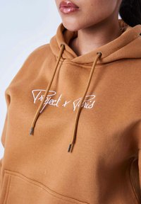 Bruine hoodie van zacht materiaal, met een voorkruiszak, trekkoorden met zilveren punten en witte geborduurde tekst.
