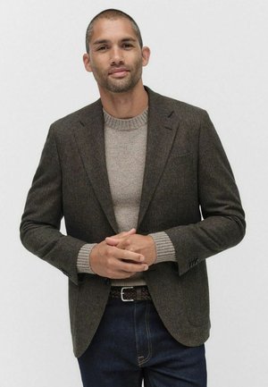 Brun tweedblazer med vevpolar, tillsammans med en ljusbeige tröja. Mörka jeans och ett flätat bälte kompletterar outfiten.