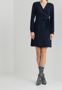 Námořnické modré šaty se zavazováním v pase, dlouhými rukávy a délkou ke kolenům. Outfit doplňují vzorované černobílé boty.