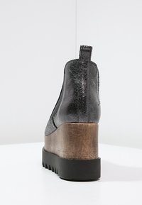 Musta nilkkuri, jossa on rakenneleather, puinen paksu korko ja kumipohja; varustettu vetotarralla ja sahalaitakuviolla pohjassa.