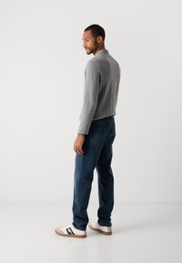Man standing sideways wearing a gray long-sleeve shirt, dark blue jeans, and white sneakers with black stripes on a white background.