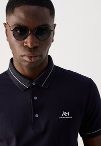 Zwarte polo met een gestructureerde kraag, lichtblauwe knopen en witte accenten; model draagt ronde zonnebril, met een geconcentreerde uitstraling.