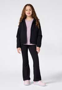 Jeune fille aux cheveux bouclés portant une veste noire zippée, un t-shirt lavande, un pantalon noir et des baskets roses et blanches, se tenant sur un fond blanc.