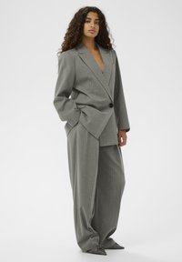 Femeie cu păr lung și ondulat, îmbrăcată într-un costum gri cu dungi verticale, de dimensiuni ample, cu pantaloni evazați și pantofi ascuțiți, stând în fața unui fundal alb.