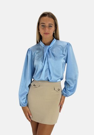 Blusa di satin blu chiaro con scollatura a fiocco e maniche lunghe a sbuffo, abbinata a una mini gonna beige con bottoni decorativi.