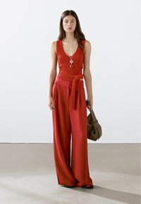 Röd stickad topp med djup v-ringning och matchande vida byxor med knyt i midjan. Modellen håller en brun handväska och står framför en enfärgad bakgrund.