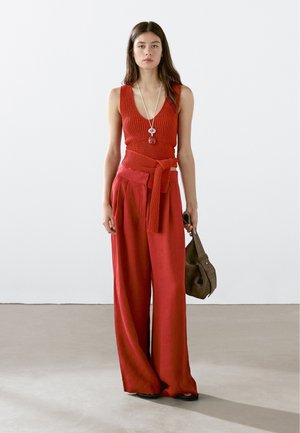 Rød striktop med dyb v-hals og matchende bukser med bred ben, der har en bundet talje. Modellen holder en brun håndtaske og står foran en ensfarvet baggrund.