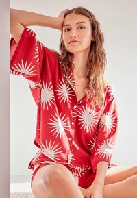 Ensemble de pyjama rouge avec un motif de formes blanches en rayons de soleil ; manches courtes, devant à boutons, et short à coupe décontractée.