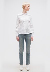 Άτομο με κοντά ξανθά μαλλιά που φοράει λευκό πουκάμισο με κουμπιά, ανοιχτό μπλε τζιν και λευκά αθλητικά παπούτσια, όρθιο μπροστά σε απλό λευκό φόντο.