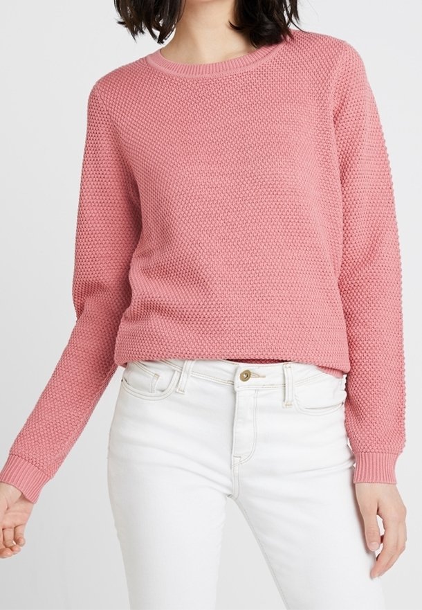 Maglione rosa lavorato a maglia con un design testurizzato, maniche lunghe e un collo a costine. Abbinato a jeans bianchi e un dettaglio con bottoni dorati.
