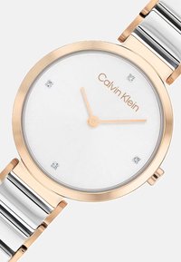 Montre Calvin Klein avec un cadran rond blanc, des accents dorés et un bracelet en métal argenté. Présente quatre index en pierre et des aiguilles minimalistes.