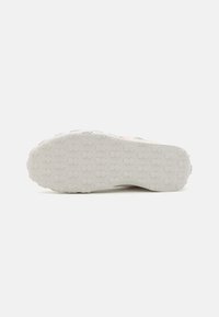 Suola di scarpa bianca con un motivo di presa testurizzato, caratterizzata da forme a diamante e freccia in rilievo per la trazione. Materiale in gomma liscia, forma arrotondata.