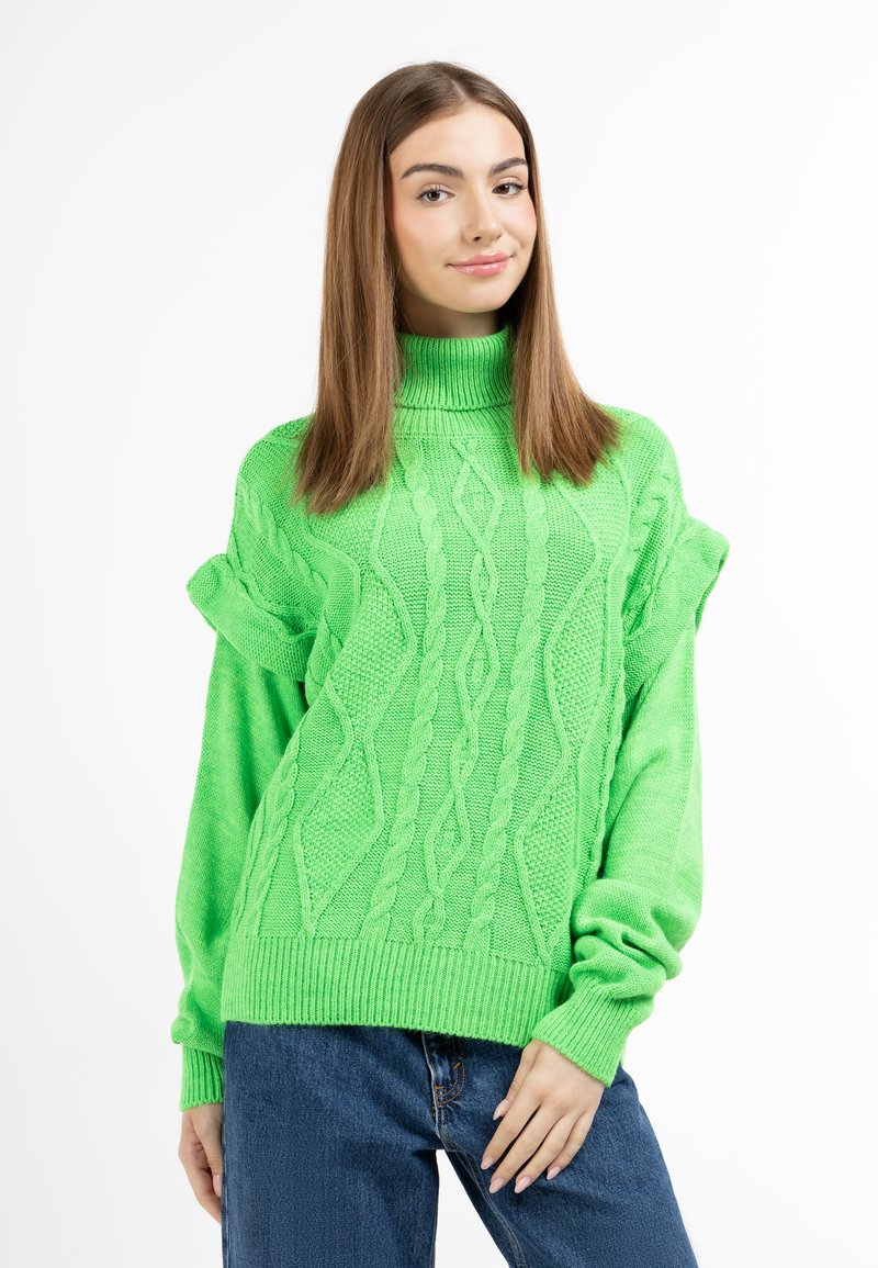 Pulover tricotat verde cu guler înalt și modele texturate de cablu. Prezintă mâneci răsucite și margine cu nervuri. Combinat cu blugi albaștri.