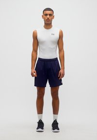 Νέος άνδρας όρθιος, ντυμένος με λευκό αμάνικο αθλητικό μπλουζάκι, μπλε σκούρο σορτς, λευκές κάλτσες και μαύρα παπούτσια για τρέξιμο, μπροστά σε λευκό απλό φόντο.