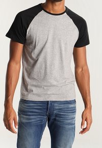 Szara koszulka z krótkim rękawem i czarnymi rękawami raglanowymi. Materiał wydaje się miękki, ma luźny krój oraz okrągły dekolt. Nosi ją z niebieskimi dżinsami.