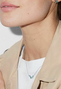 Collar de plata con piedras preciosas multicolores, que presenta una delicada cadena y tres elementos decorativos. Tono de piel claro y chaqueta beige presentes.