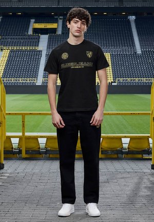 Giovane uomo in piedi sul corridoio dello stadio con indosso una t-shirt e pantaloni neri Guess Jeans con scarpe da ginnastica bianche, spalti vuoti sullo sfondo.