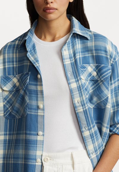 Camicia a quadri blu e crema con taschini abbottonati, realizzata in un tessuto con una superficie leggermente strutturata, indossata sopra una t-shirt bianca a coste.