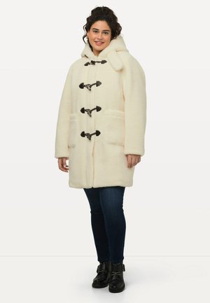 Cappotto toggle di colore crema, morbido e con cappuccio, caratterizzato da chiusure toggle nere e due tasche frontali. Abbinato a jeans scuri e stivali.