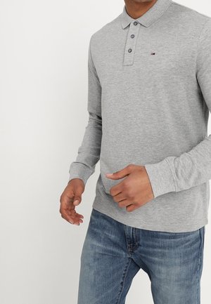 Polo gris à manches longues avec patte de boutonnage à trois boutons, poignets côtelés et petit détail de logo sur la poitrine, associé à un jean bleu.