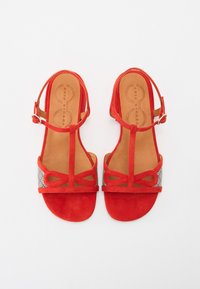 Sandali rossi in suede con design a T, caratterizzati da un pannello decorativo a motivi nere e bianche e chiusura con fibbia alla caviglia.