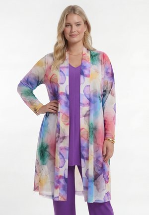 Frau trägt eine lange, farbenfrohe, transparente Blumenstrickjacke über lila Top und Hose, posiert mit Hand in der Hüfte und leichtem Make-up.