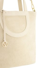 Beige ruskinds tote taske med læderdetaljer, der har en cirkulær frontlomme, glatte håndtag og en guld logo charme hængende.