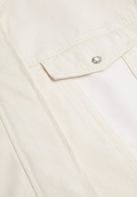 Krémová košile s vertikální žebrovanou texturou, vybavená patentním knoflíkem na náprsní kapse a uvolněným střihem.